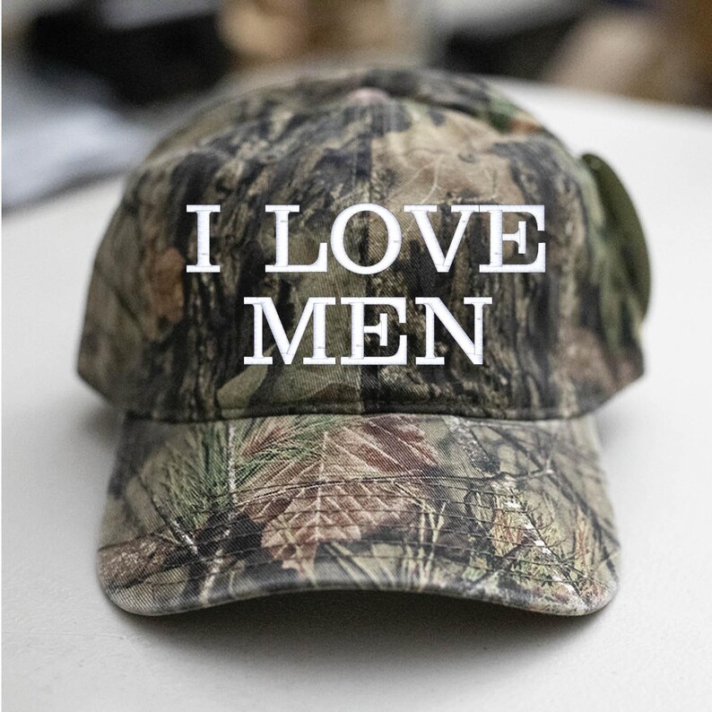 Op de afbeelding: Een camouflage baseballpet met de tekst "I LOVE MEN" in witte letters.
