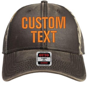 Könnte beinhalten: Eine braune und schwarze Baseballkappe mit Tarnmuster und einem orangefarbenen gestickten Text, der "CUSTOM TEXT" lautet. Die Kappe hat ein weißes Etikett an der Seite, auf dem "OTTO" in Rot und "CLASSIC FIT OSFM CAMO MAXIMUM COMFORT" in Schwarz steht.