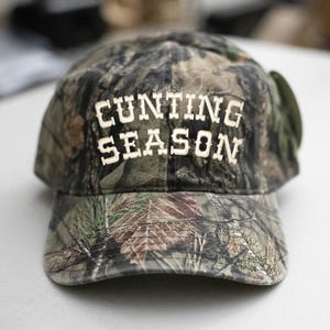 May include: A camouflage baseball cap with the words "CUNTING SEASON" in white block letters across the front. The cap has a curved peak and a classic six-panel design, suitable for outdoor pursuits.