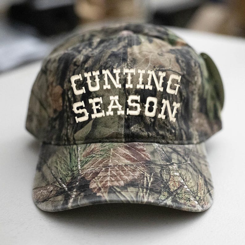 May include: A camouflage baseball cap with the words "CUNTING SEASON" in white block letters across the front. The cap has a curved peak and a classic six-panel design, suitable for outdoor pursuits.