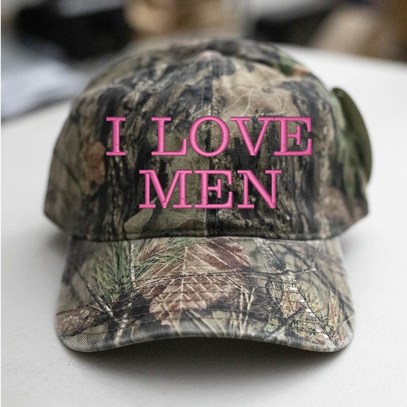 Op de afbeelding: Een camouflage baseballpet met de woorden "I LOVE MEN" geborduurd in roze.