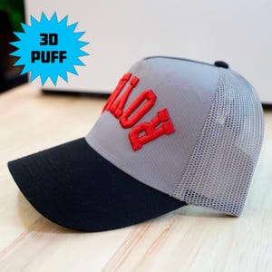 Puede incluir: Una gorra de camionero gris y negra con bordado rojo en relieve 3D en el panel frontal. La gorra tiene una visera negra sólida, una corona frontal gris y una parte trasera de malla gris. Un gráfico azul muestra el texto 3D PUFF.