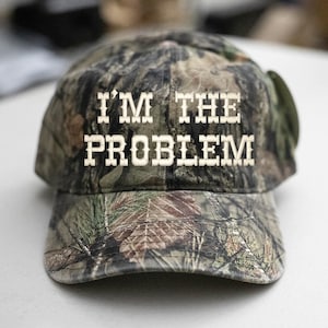Puede incluir: Gorra de béisbol de camuflaje con el texto "I'M THE PROBLEM" en letras blancas. La gorra tiene una visera curva y un diseño clásico de seis paneles. La tela tiene un patrón de color verde, marrón y beige.
