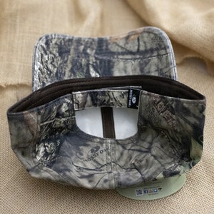 May include: A brown and green camouflage baseball cap with a Mossy Oak logo on the front and back. The cap has a brown adjustable strap at the back.