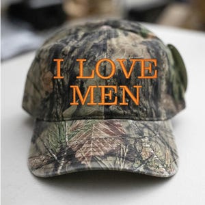 Op de afbeelding: Een camouflage baseballpet met de tekst "I LOVE MEN" geborduurd in oranje.