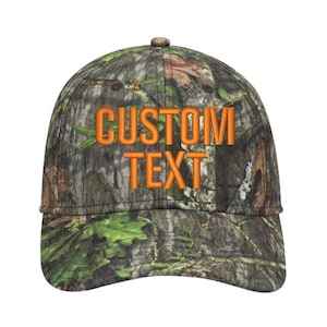 May include: A camouflage baseball cap with the words "CUSTOM TEXT" embroidered in orange. The cap features a curved brim and a button on top. The camouflage pattern includes shades of green, brown, and tan.