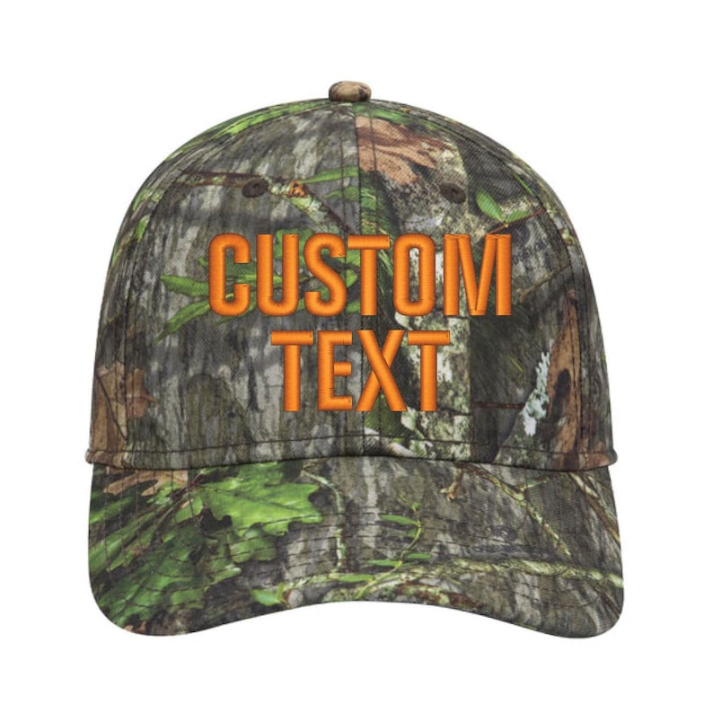 May include: A camouflage baseball cap with the words "CUSTOM TEXT" embroidered in orange. The cap features a curved brim and a button on top. The camouflage pattern includes shades of green, brown, and tan.