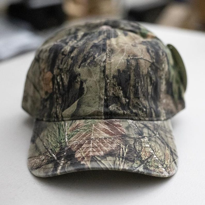 May include: A camouflage baseball cap in shades of green, brown, and tan. The cap features a curved brim and a structured crown. The fabric has a detailed pattern designed to blend into a woodland environment. The cap is suitable for outdoor activities.