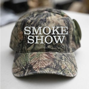 May include: A camouflage baseball cap with the text "SMOKE SHOW" in white lettering.