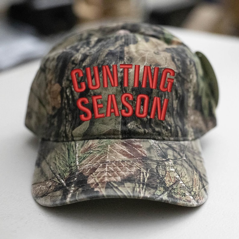 May include: Camouflage baseball cap with the embroidered text "CUNTING SEASON" in red. The cap has a curved peak and a classic six-panel design. The hat is made of a durable fabric and is suitable for outdoor activities.