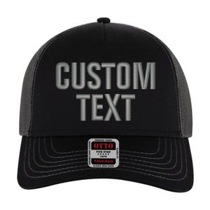 Puede incluir: Gorra de camionero negra y gris con texto bordado en gris que dice "CUSTOM TEXT". La gorra tiene una parte trasera de malla negra y una correa ajustable negra.