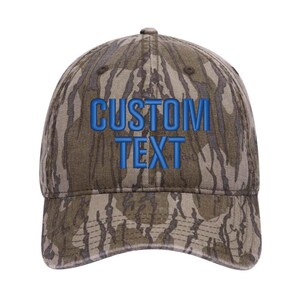 Puede incluir: Una gorra de b&eacute;isbol de camuflaje con texto bordado en azul que dice "CUSTOM TEXT".