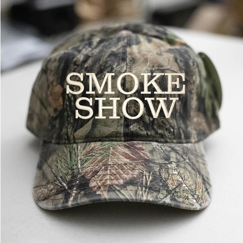 May include: A camouflage baseball cap with the words "SMOKE SHOW" embroidered in white on the front.