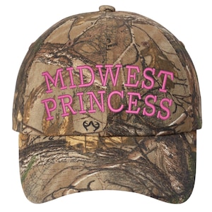 Puede incluir: Gorra de béisbol de camuflaje con las palabras "MIDWEST PRINCESS" bordadas en rosa. La gorra tiene una visera curva y un logotipo de Realtree. El sombrero está diseñado para actividades al aire libre y uso casual.