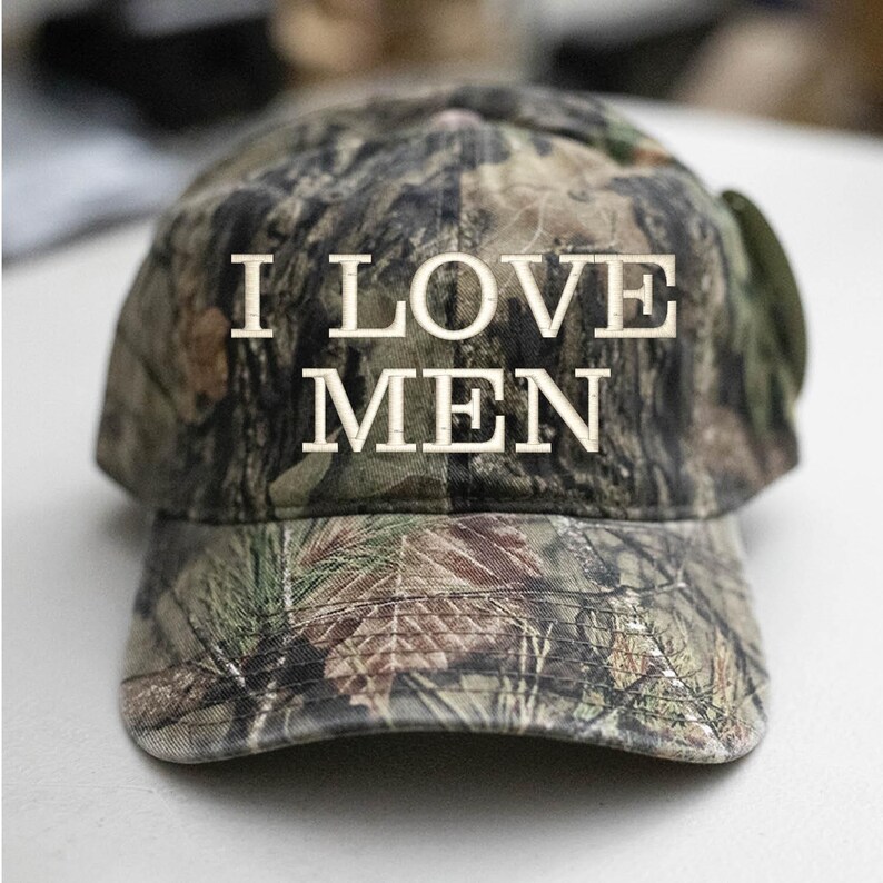 Op de afbeelding: Een camouflage baseballpet met de tekst "I LOVE MEN" geborduurd in wit.
