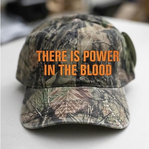 Può includere: Un cappello da baseball mimetico con il testo "THERE IS POWER IN THE BLOOD" ricamato in arancione.
