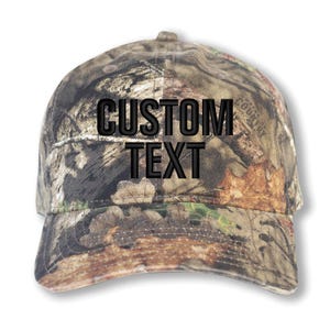May include: A camouflage baseball cap with the words "CUSTOM TEXT" embroidered in black across the front. The cap features a curved brim and a structured crown. The camouflage pattern includes shades of brown, green, and tan.