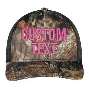 May include: A black and brown camouflage trucker hat with a pink embroidered "CUSTOM TEXT" on the front.
