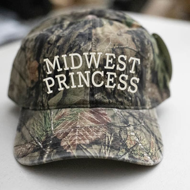 May include: A camouflage baseball cap with the text "MIDWEST PRINCESS" embroidered in white on the front.