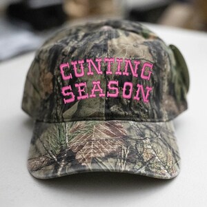 May include: A camouflage baseball cap with the words "CUNTING SEASON" embroidered in bright pink. The cap features a curved brim and a classic six-panel design, suitable for outdoor activities.