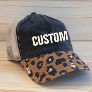 May include: A black baseball cap with a mesh back and a brown leopard print brim. The front panel features the word CUSTOM embroidered in white capital letters. The cap has a structured crown and a curved visor with visible stitching details.