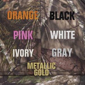 Op de afbeelding: Een close-up van een camouflage stof met verschillende gekleurde borduursels die de woorden "ORANGE", "BLACK", "PINK", "WHITE", "IVORY", "GRAY" en "METALLIC GOLD" tonen.