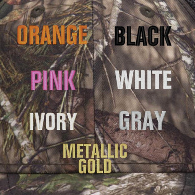 Op de afbeelding: Een close-up van een camouflage stof met verschillende gekleurde borduursels die de woorden "ORANGE", "BLACK", "PINK", "WHITE", "IVORY", "GRAY" en "METALLIC GOLD" tonen.