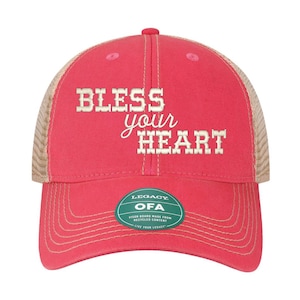 Może przedstawiać: Czerwono-brązowa czapka trucker z haftem "Bless your heart" w kolorze białym z przodu. Czapka ma zieloną metkę z tekstem "Legacy OFA Visor Board Made from Recycled Content Live Your Legacy".
