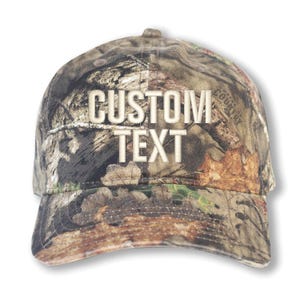 May include: A camouflage baseball cap with the words "CUSTOM TEXT" embroidered in white. The cap features a curved brim and a classic six-panel design. The camouflage pattern includes shades of brown, green, and tan.