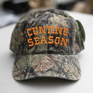 May include: A camouflage baseball cap with the words "CUNTING SEASON" embroidered in orange thread. The cap has a curved peak and a classic six-panel design, suitable for outdoor activities.