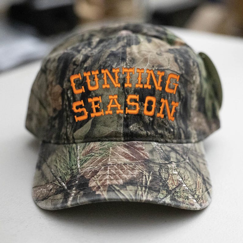 May include: A camouflage baseball cap with the words "CUNTING SEASON" embroidered in orange thread. The cap has a curved peak and a classic six-panel design, suitable for outdoor activities.