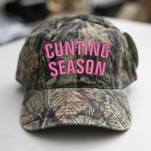 May include: A camouflage baseball cap with the words "CUNTING SEASON" embroidered in bright pink. The hat has a curved peak and a classic six-panel design, suitable for outdoor activities.