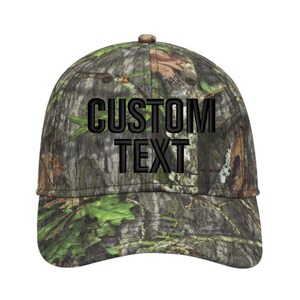 May include: A camouflage baseball cap with the words "CUSTOM TEXT" embroidered in black. The cap features a curved brim and a button on top. The camouflage pattern includes shades of green, brown, and tan.