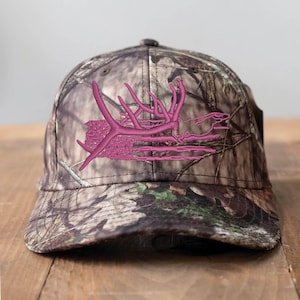May include: A camouflage baseball cap with a pink embroidered antler design. The cap features a curved brim and a classic six-panel construction. The hat is made of a durable fabric and is suitable for outdoor activities.