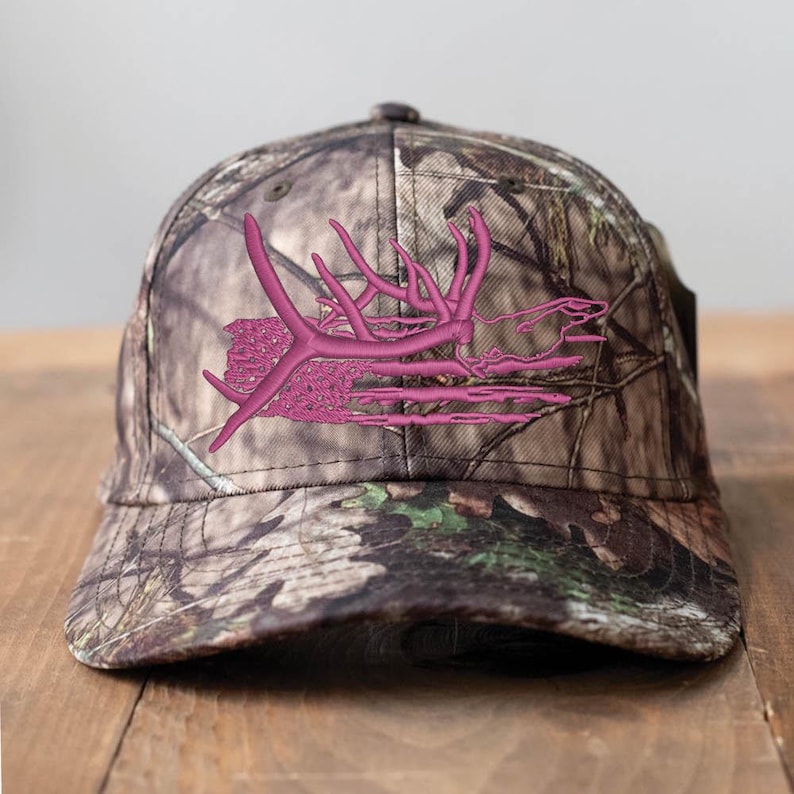 May include: A camouflage baseball cap with a pink embroidered antler design. The cap features a curved brim and a classic six-panel construction. The hat is made of a durable fabric and is suitable for outdoor activities.