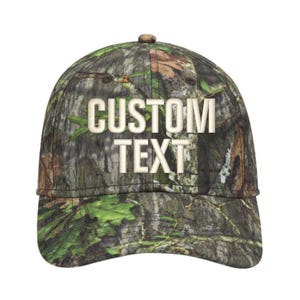 May include: A camouflage baseball cap with the words "CUSTOM TEXT" embroidered in a light beige color. The cap features a curved brim and a button on top. The camouflage pattern includes shades of green, brown, and tan.