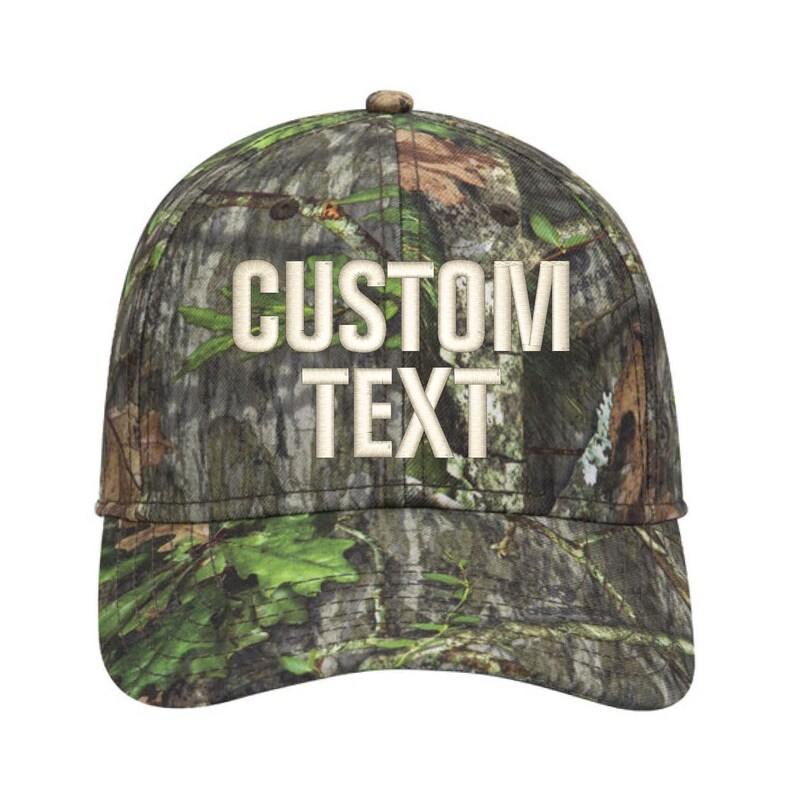 May include: A camouflage baseball cap with the words "CUSTOM TEXT" embroidered in a light beige color. The cap features a curved brim and a button on top. The camouflage pattern includes shades of green, brown, and tan.
