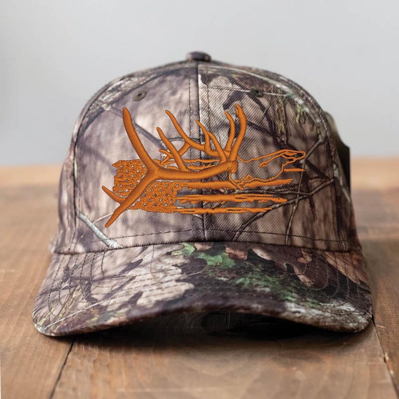 May include: A camouflage baseball cap with an embroidered orange antler design. The cap features a curved brim and a classic six-panel construction. The hat is designed for outdoor enthusiasts and hunters.