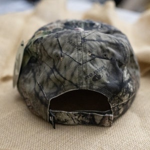 May include: A camouflage baseball cap with the words "Mossy Oak" printed on the back. The cap is made of a fabric with a pattern of green, brown, and gray, and has a black adjustable strap.