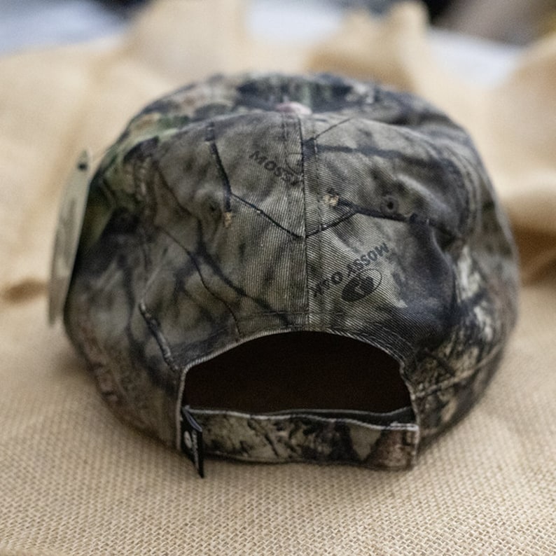 May include: A camouflage baseball cap with the words "Mossy Oak" printed on the back. The cap is made of a fabric with a pattern of green, brown, and gray, and has a black adjustable strap.