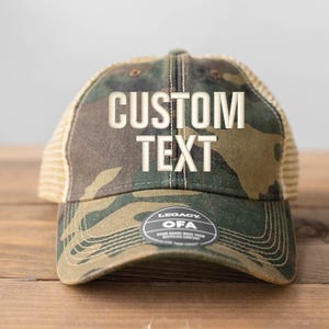 May include: A camouflage trucker hat with a tan mesh back. The hat has the words "CUSTOM TEXT" embroidered on the front in white.