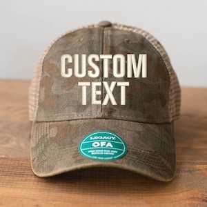 May include: A camouflage-patterned baseball cap with the words "CUSTOM TEXT" embroidered in white. The cap has a mesh back and a green and white sticker on the brim. The hat is on a wooden surface.