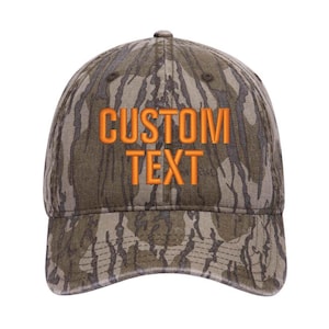 Puede incluir: Una gorra de b&eacute;isbol de camuflaje con el texto "CUSTOM TEXT" bordado en naranja.