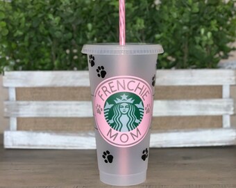May include: A clear plastic reusable cup with a pink lid and a pink and white striped straw. The cup has a black and white paw print design and a green and white logo that says "Frenchie Mom".