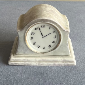 Pode incluir: Um relógio de lareira de cerâmica branca com marcadores de hora em algarismos romanos pretos e ponteiros pretos. O relógio tem um mostrador branco e uma moldura dourada.