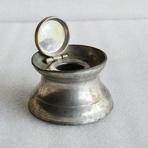 Puede incluir: Un tintero de metal vintage con una base ancha y acampanada y una abertura circular. La pieza cuenta con una tapa circular con bisagras que se encuentra en posición abierta, revelando un interior liso. La superficie metálica tiene un acabado mate.