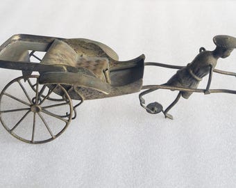 May include: A metallic, brass-toned miniature rickshaw figurine featuring a seated carriage area with spoked wheels and a figure wearing a conical hat positioned at the front, appearing to pull the vehicle by two long, thin metal handles.