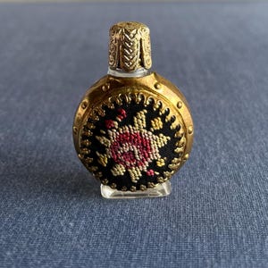 Puede incluir: Un frasco de perfume vintage ornamentado, con un exterior de metal dorado. El frasco presenta un panel de tela negra con un diseño floral en rojo, amarillo y blanco. El frasco se apoya sobre una base transparente y rectangular.