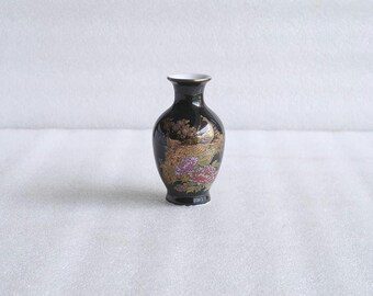 May include: A small black ceramic vase with a narrow neck and flared rim. The body features a detailed gold-colored peacock illustration surrounded by vibrant red and purple floral accents, finished with a glossy surface and a white interior rim.