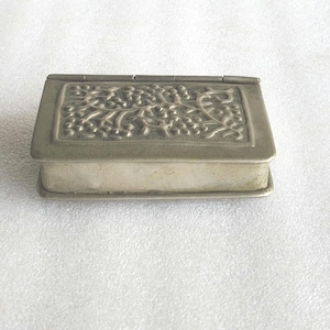 Puede incluir: Una caja rectangular de metal plateado con tapa abatible. La superficie superior presenta un patrón en relieve texturizado que representa una enredadera con racimos de bayas y hojas, enmarcado por un borde liso y sencillo.
